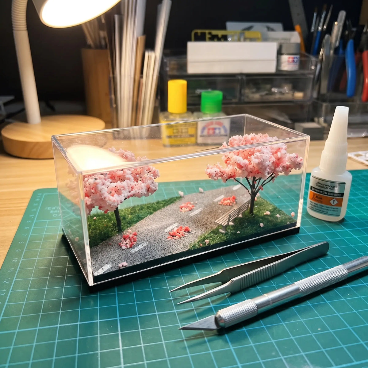 A clear acrylic diorama box containing a miniature road, green grass, and pink cherry blossom trees, displayed on a green cutting mat with tweezers, a craft knife, and glue nearby.